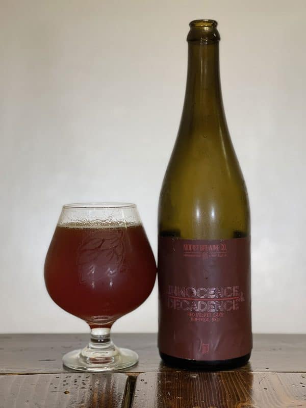 Snifter glass of beer sitting next to a bottle of Modist Brewing Innocence & Decadence Red Velvet Cake Imperial Red.