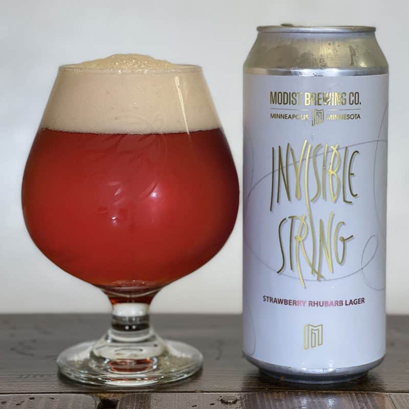 Glass filled with beer sitting next to a can of Modist Brewing Invisible String Strawberry Rhubarb Lager.