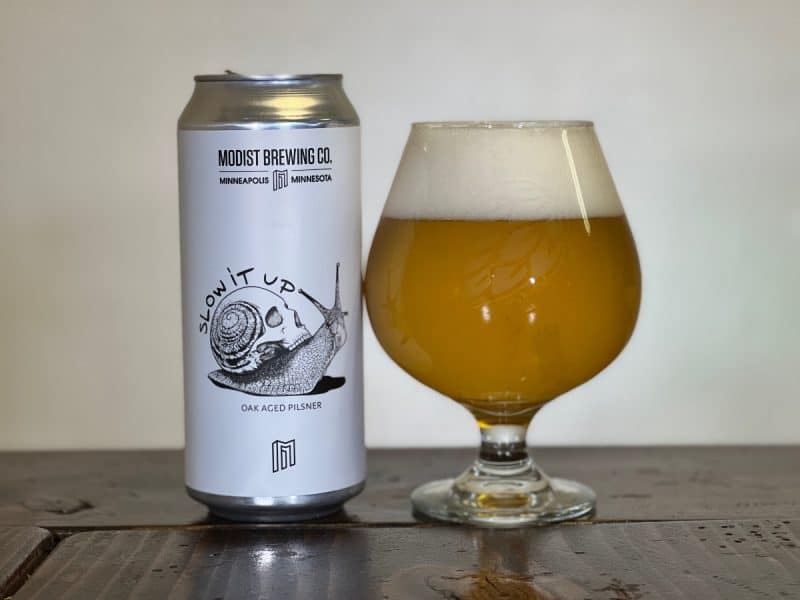 Glass of beer next to a can of Modist Brewing Slow It Up Oak-Aged Pilsner.