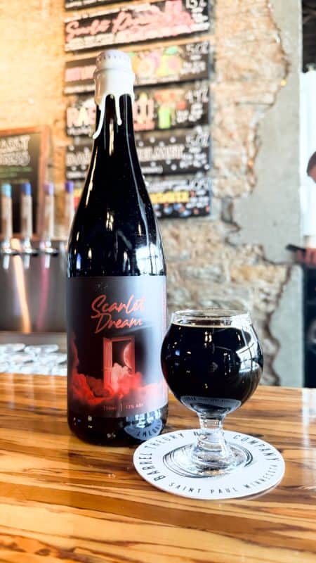 Bottle of Barrel Theory Brewing Scarlet Dream sitting next to a small glass of deep black liquid at the bar at Barrel Theory Brewing.