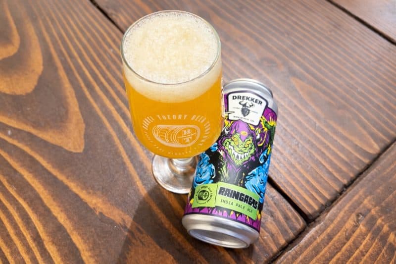 Can of Drekker Brewing. and Barrel Theory Raingasms laying next to a glass filled with beer, on a wooden table.