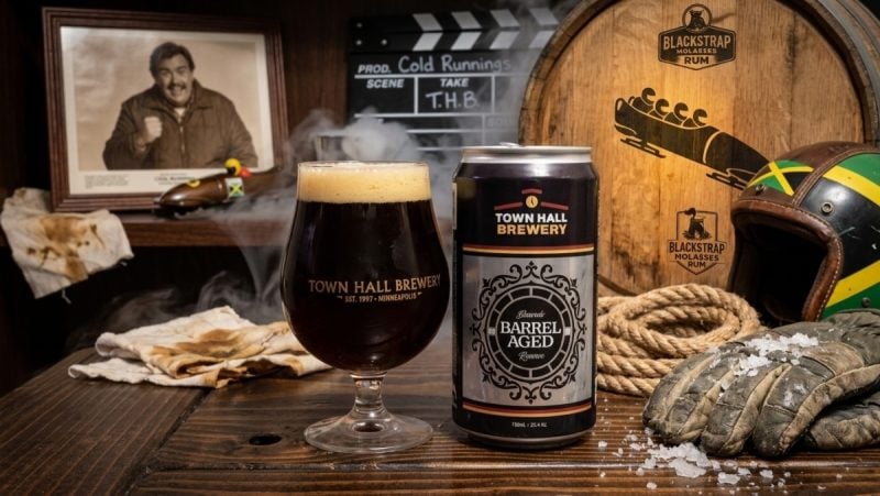 A glass and crowler of Town Hall Brewery Cold Runnings with Jamaican bobsled memorabilia and a photo of John Candy.