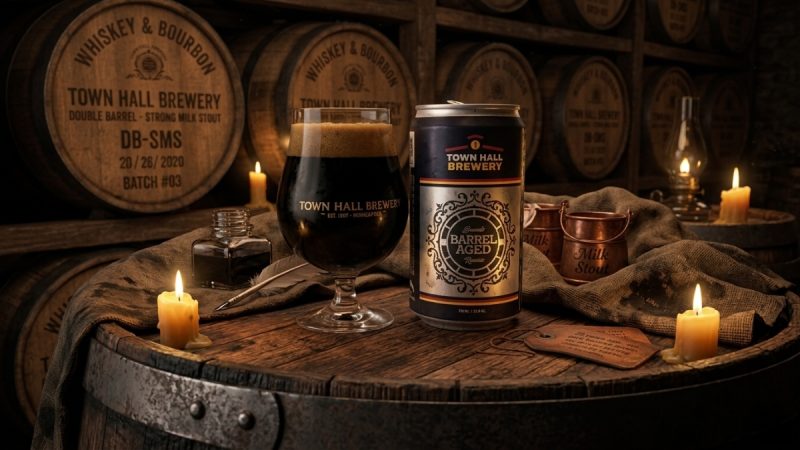 Glass and crowler of Town Hall Brewery Double Barrel Xtra Milk Stout sitting on a barrel in a barrel room.